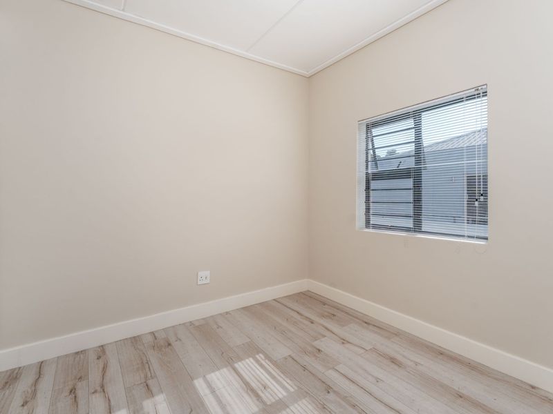 A bright, empty bedroom featuring modern laminate flooring, neutral walls, and a built-in cupboard with aluminium window frames and venetian blinds.