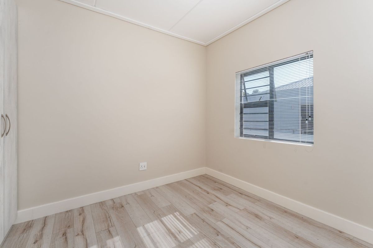 A bright, empty bedroom featuring modern laminate flooring, neutral walls, and a built-in cupboard with aluminium window frames and venetian blinds.