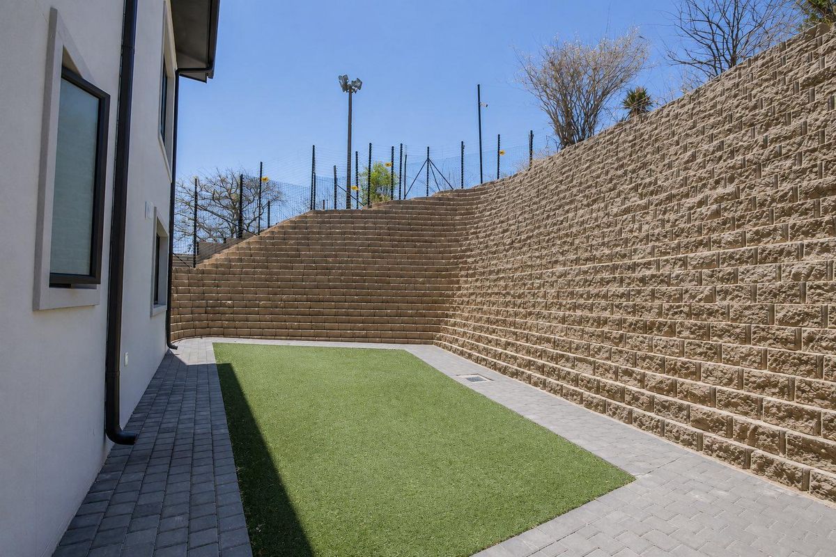 A low-maintenance rear courtyard featuring a large tiered retaining wall, artificial turf lawn, and paved perimeter walkway.