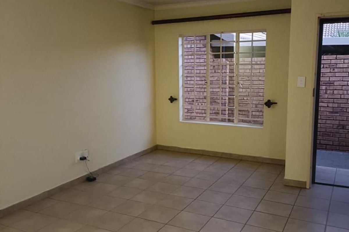 An empty, neutrally painted living room featuring tiled flooring, a window with security bars, and direct access to an exterior area.