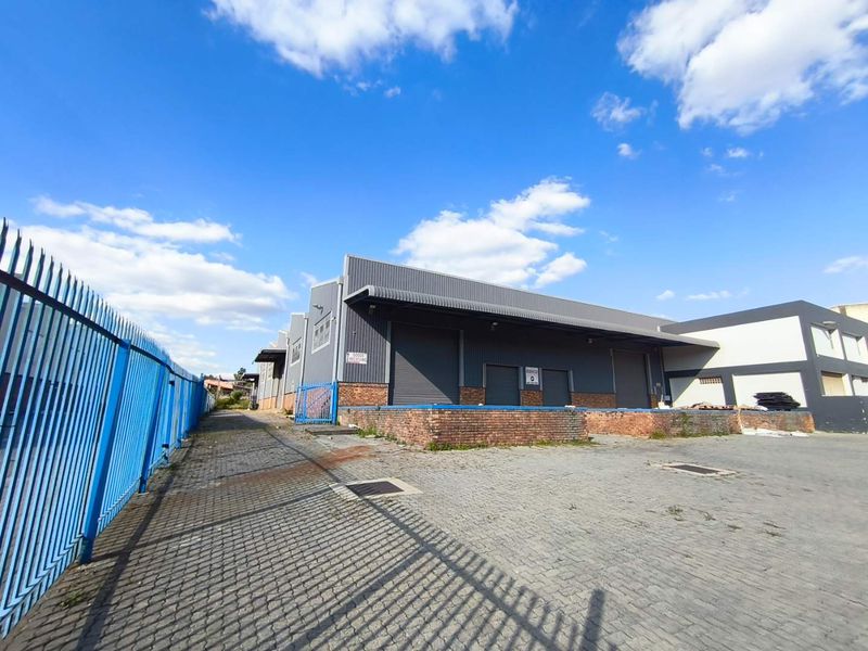 Industrial warehouse exterior featuring a loading dock, multiple roller shutter doors, and a paved yard area enclosed by security fencing.