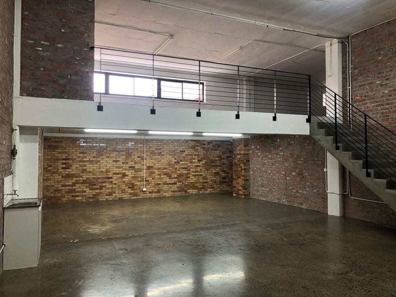 Spacious industrial-style open plan area featuring exposed brick walls, polished concrete floors, a mezzanine level with metal staircase, and basic utility sink.