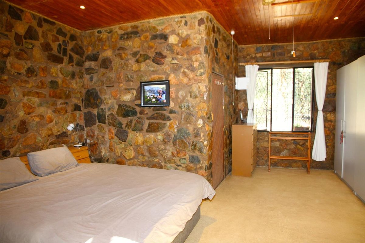 A rustic bedroom featuring a prominent natural stone feature wall, timber-clad ceiling, and large window providing natural light.