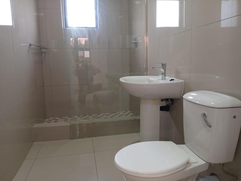 A clean, modern bathroom featuring a pedestal basin, a white toilet, and a glass-enclosed shower with neutral wall and floor tiling.