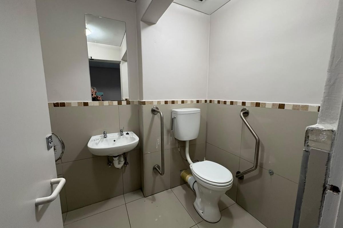 A functional office bathroom featuring a wall-mounted basin, toilet, and accessibility grab rails, with neutral tiling throughout.