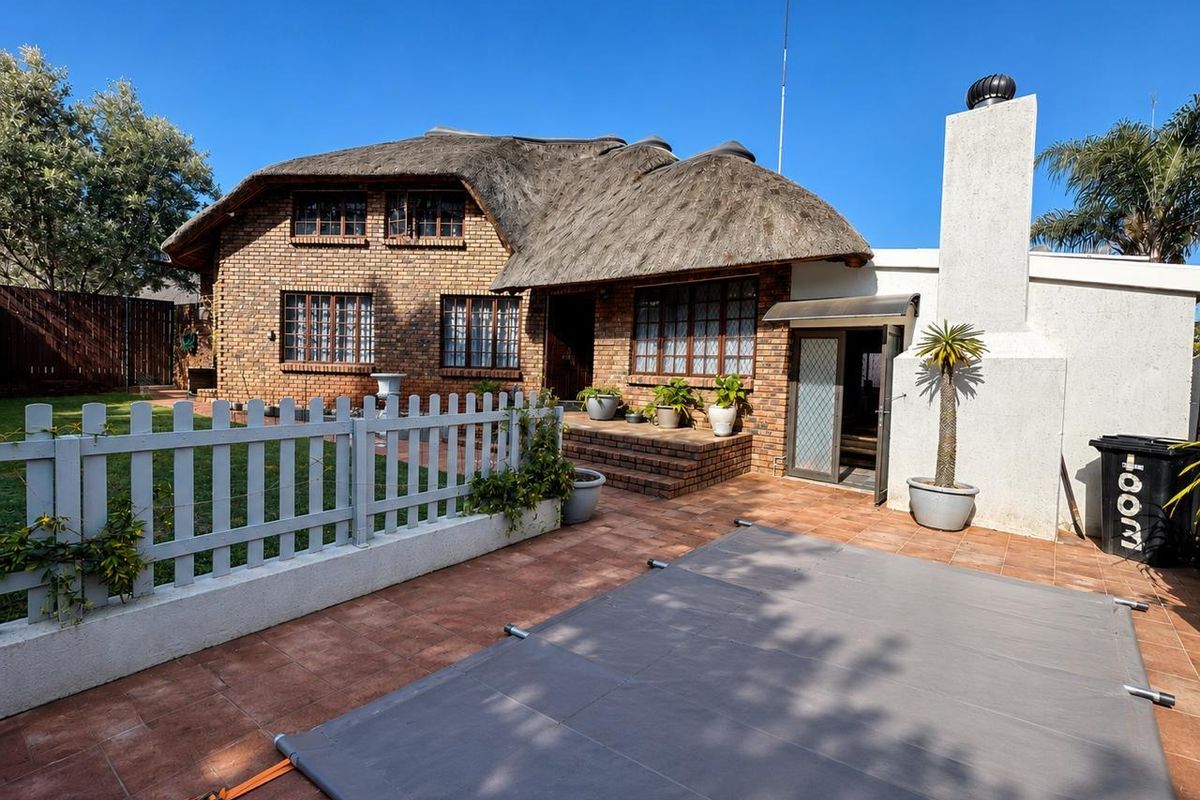 Rear view of a face brick house with a traditional thatch roof, featuring a tiled patio area and a swimming pool with a cover.