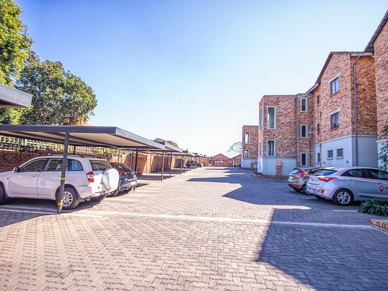Exterior view of the townhouse complex featuring a paved parking area with covered carports and multi-story brick residential buildings.