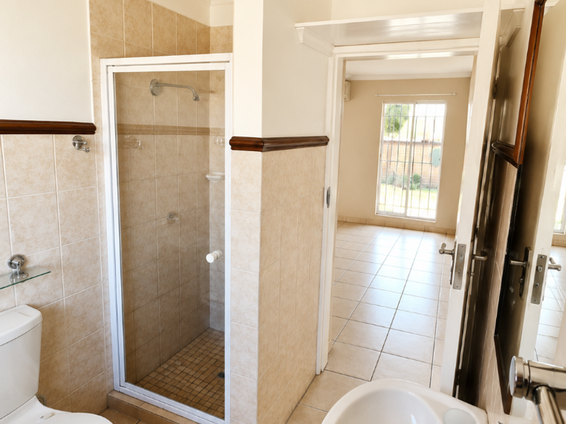 A clean, functional bathroom featuring a shower enclosure, pedestal basin, and neutral ceramic tiling, with a doorway leading to an adjacent room.