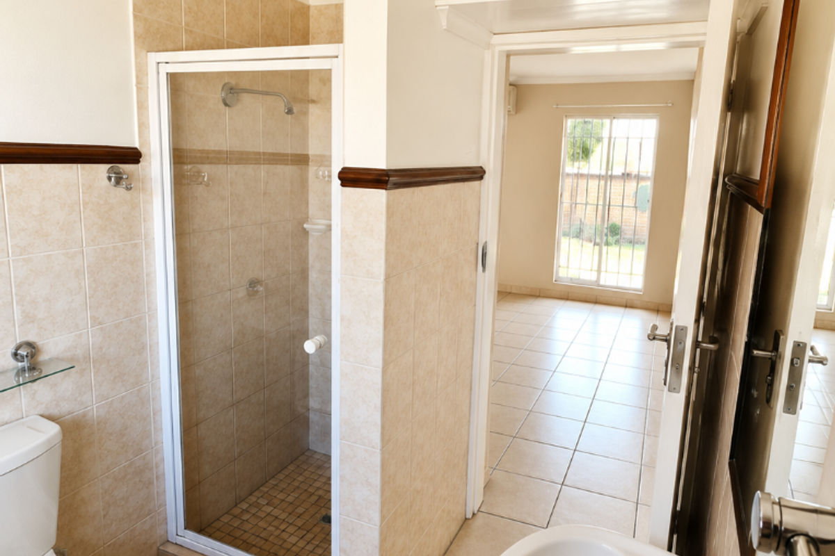A clean, functional bathroom featuring a shower enclosure, pedestal basin, and neutral ceramic tiling, with a doorway leading to an adjacent room.