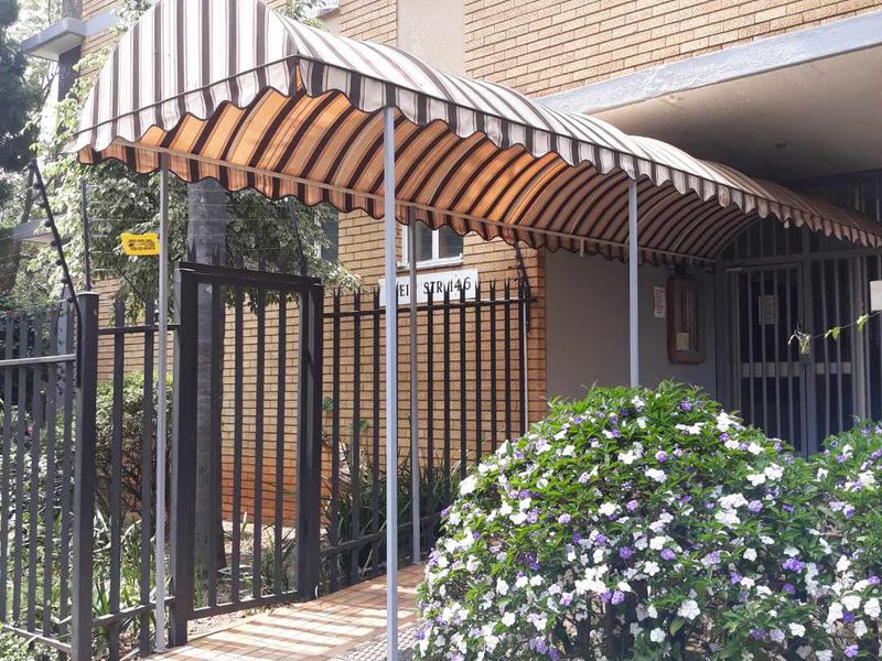 Front entrance of the apartment building featuring a striped fabric awning, paved walkway, and secure metal gate access.