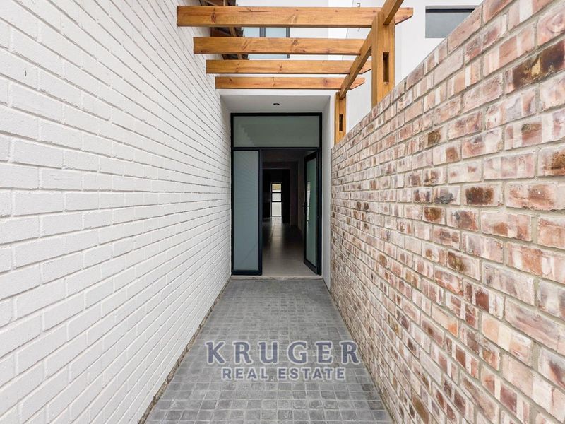 A modern entrance walkway featuring a timber pergola, contrasting painted and exposed brick walls, and durable cobblestone paving leading to a glass panel door.