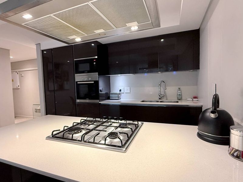 Modern kitchen featuring dark gloss cabinetry, stone countertops, a central island with a gas hob, and integrated built-in appliances.