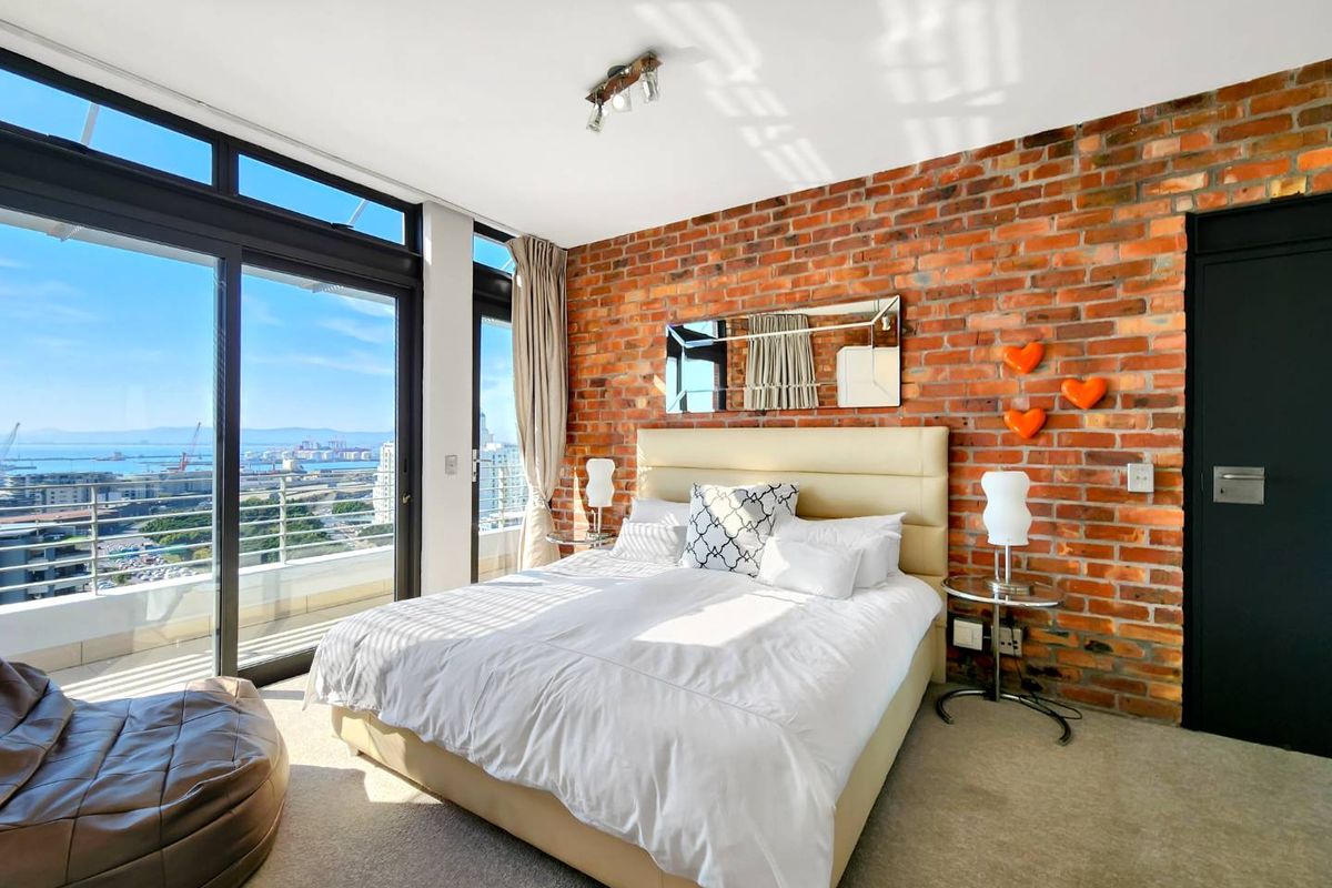 Modern bedroom featuring an exposed brick feature wall, carpeted flooring, and large sliding glass doors opening onto a balcony with expansive city and harbor views.
