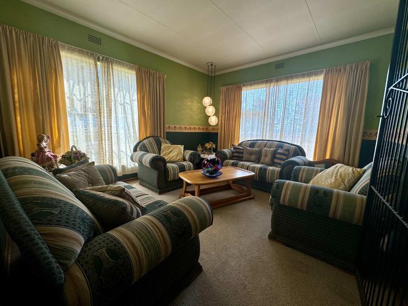 A spacious living room featuring large windows with natural light, carpeted flooring, and a central pendant light fixture.