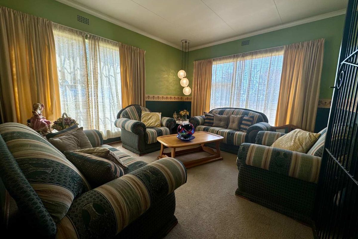 A spacious living room featuring large windows with natural light, carpeted flooring, and a central pendant light fixture.