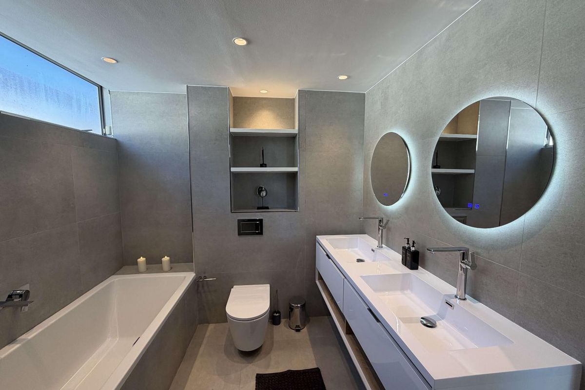 Modern bathroom featuring a double vanity, wall-hung toilet, built-in bathtub, and elegant LED backlit mirrors with contemporary grey tiling throughout.