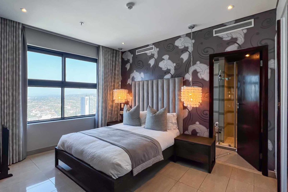 Modern bedroom featuring a large window with city views, patterned feature wall, recessed ceiling lighting, and an en-suite bathroom access.