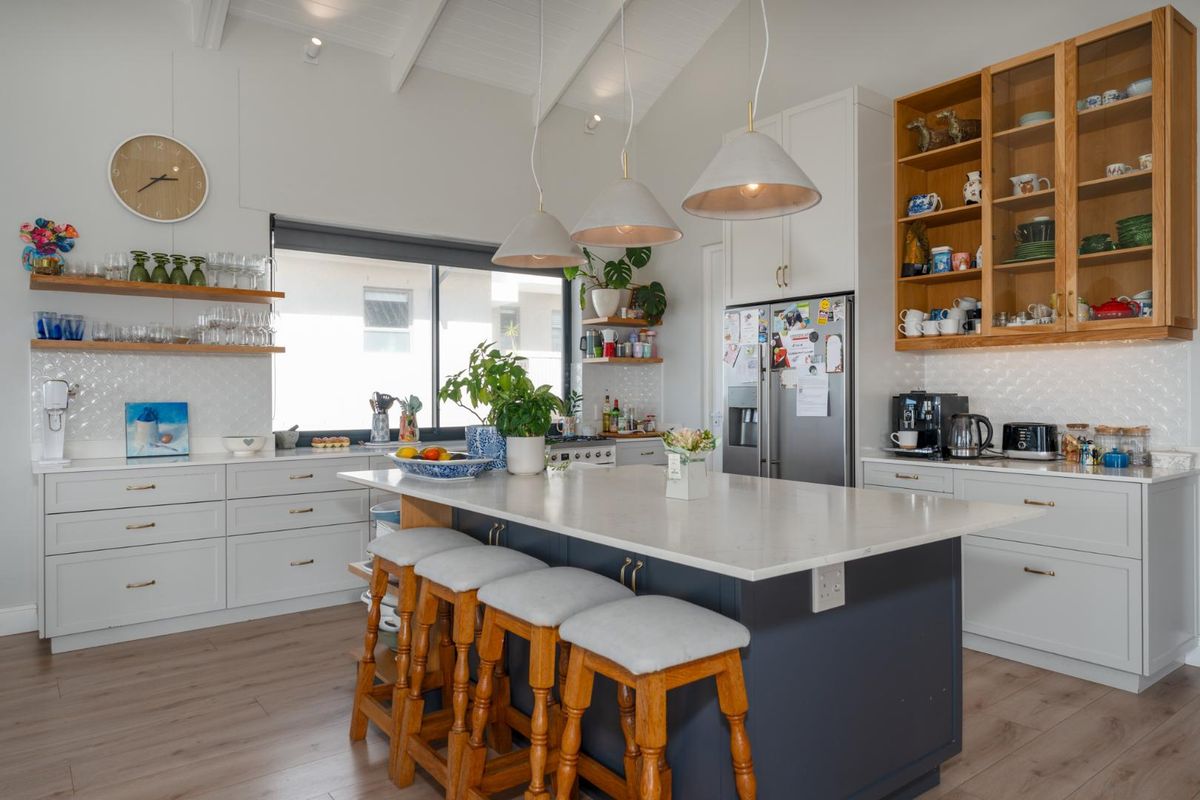 A modern, well-lit kitchen featuring a central island with stone countertops, ample built-in cabinetry, open shelving, and a high vaulted ceiling.