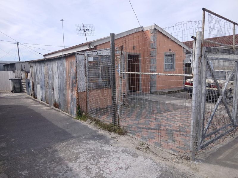 Front exterior view of a single-story house featuring a paved driveway, wire mesh perimeter fencing, and a corrugated metal side structure.