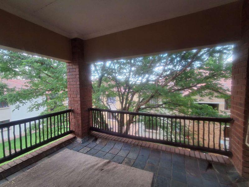 A covered balcony featuring brick pillars, a timber balustrade, and slate tile flooring, offering a view of the surrounding complex greenery.