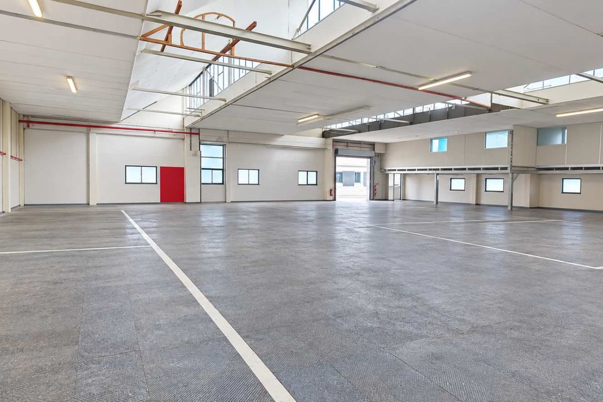 Spacious industrial warehouse featuring high ceilings, concrete flooring, ample natural light through skylights, and a large roller shutter door for easy access.