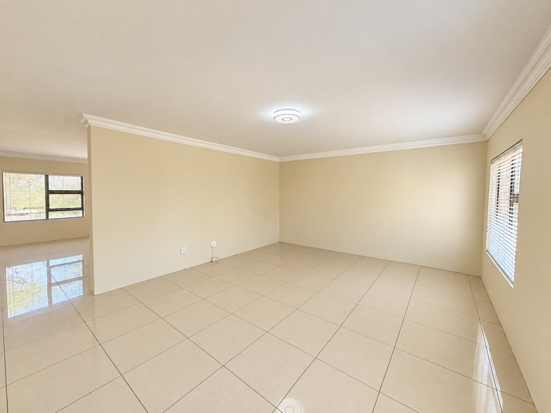 A bright, spacious, and empty living area featuring neutral tiled flooring, clean walls, ceiling cornices, and large windows with fitted blinds.