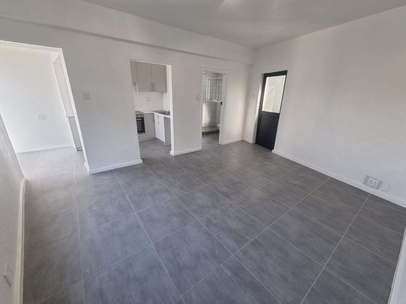 A bright, open-plan living area featuring modern grey floor tiles, leading into a compact kitchen and adjacent bathroom space.