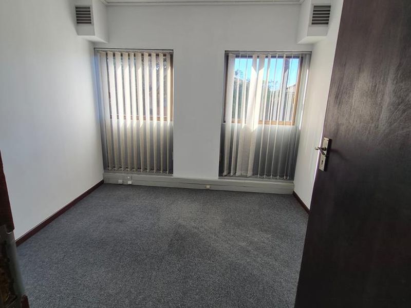 A clean, empty office space featuring grey carpet flooring, vertical window blinds, and integrated ceiling lighting, suitable for a professional workspace.
