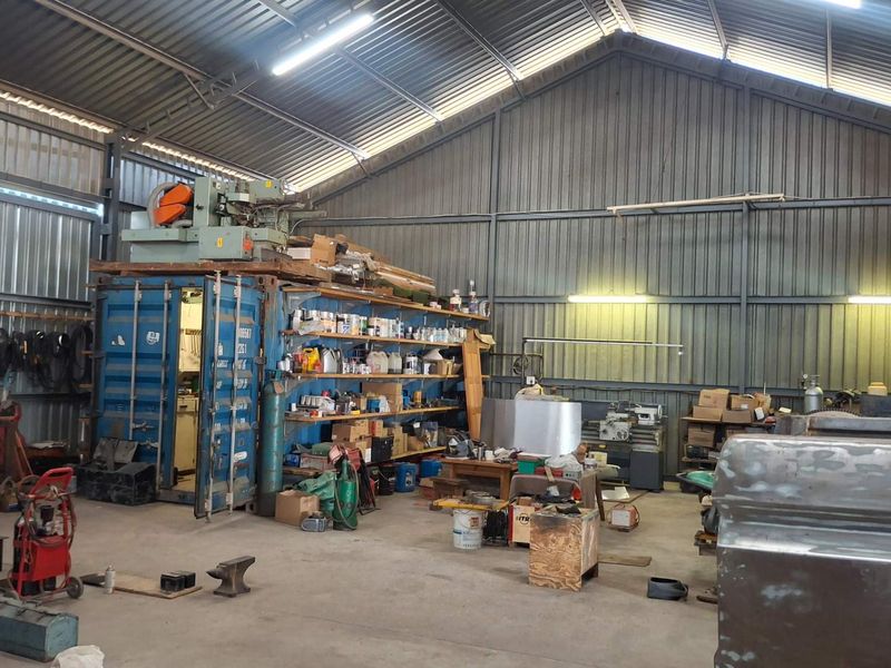 Spacious industrial workshop featuring high-clearance corrugated metal walls, exposed steel roof trusses, concrete flooring, and an integrated shipping container for secure storage.