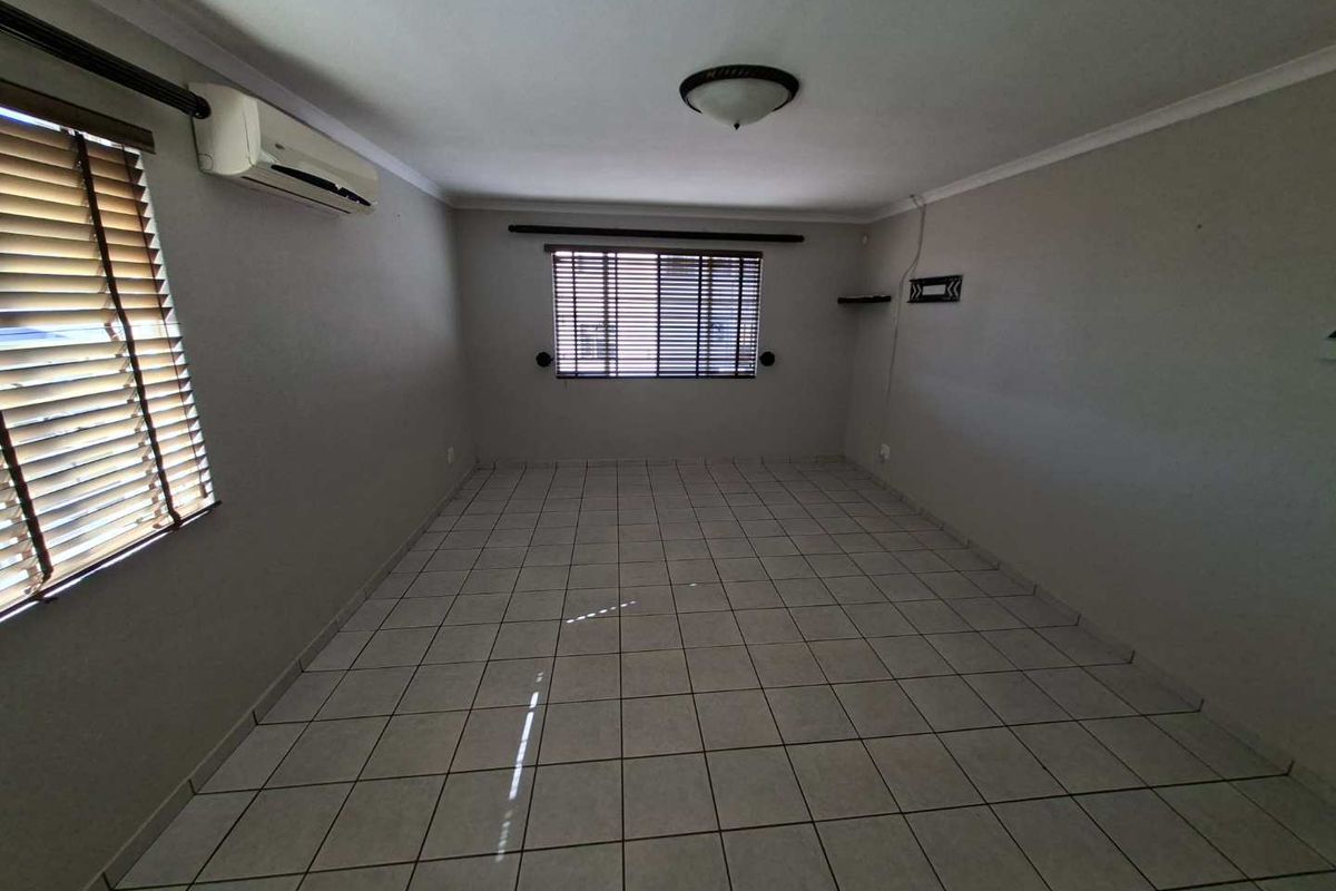A spacious, empty living area featuring neutral tiled flooring, a wall-mounted air conditioning unit, and venetian blinds on the windows.