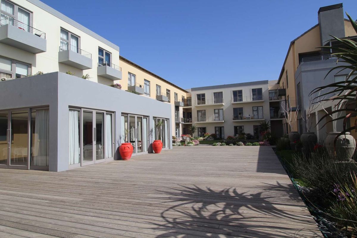 A spacious outdoor timber deck area situated within an apartment complex, featuring modern building facades and landscaped garden beds.