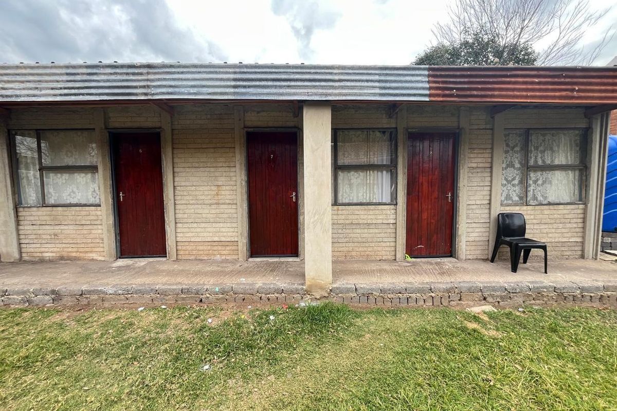 Exterior view of a row of residential units featuring brick construction, individual wooden doors, and a corrugated metal roof covering a concrete walkway.