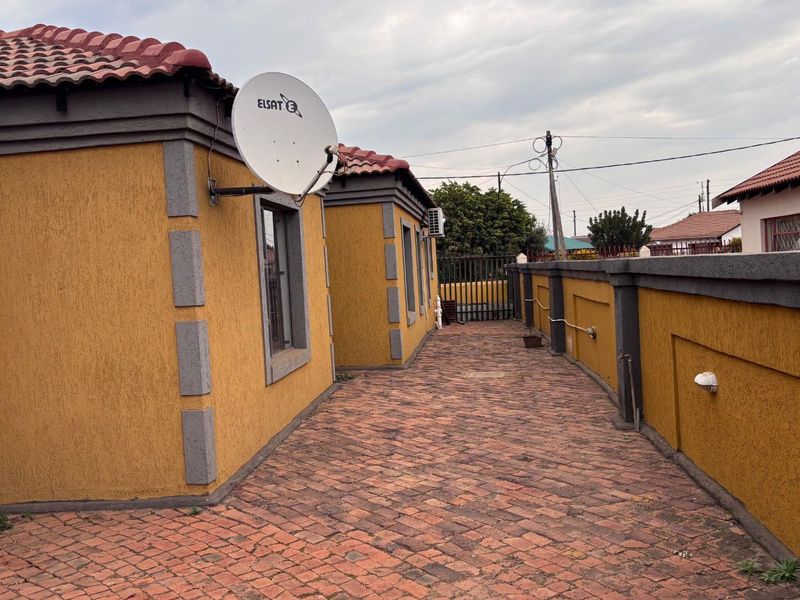 A side exterior view of the property featuring a paved walkway, textured yellow walls with grey accents, and a terracotta tiled roof.