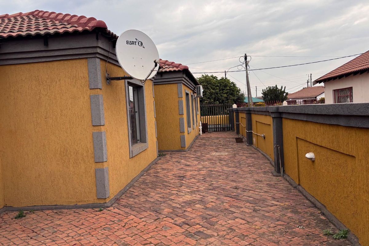 A side exterior view of the property featuring a paved walkway, textured yellow walls with grey accents, and a terracotta tiled roof.