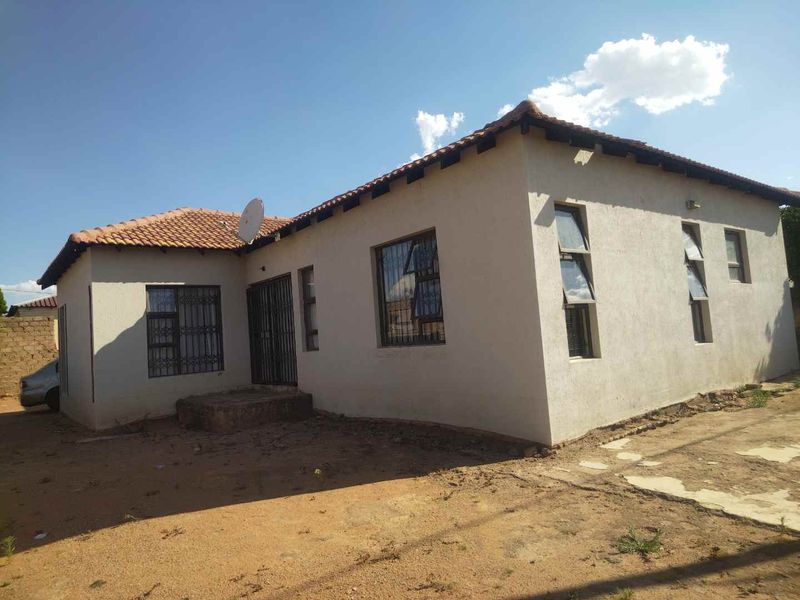 A single-story residential house featuring a terracotta tiled roof, security bars on windows, and an unpaved yard area.