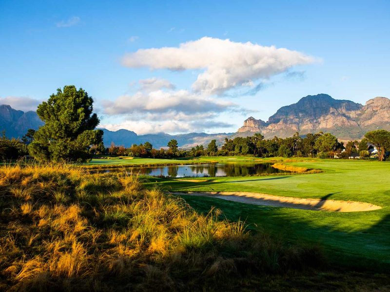 A scenic view of a well-maintained golf course featuring a water hazard, sand bunker, and mountain backdrop.