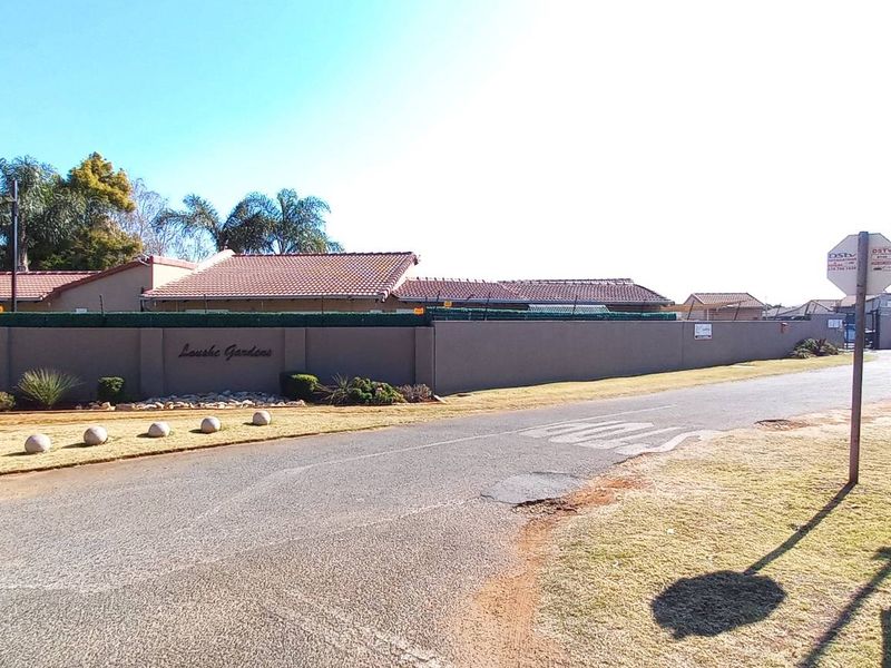Street view of the townhouse complex entrance featuring perimeter walling, security fencing, and tiled roof structures.
