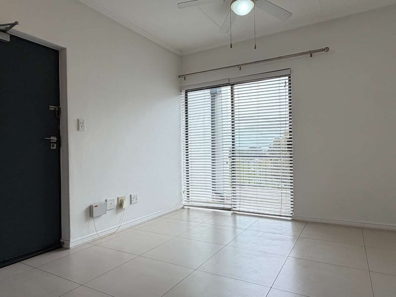 An empty, well-lit living area featuring neutral tiled flooring, a ceiling fan, and a sliding glass door with fitted venetian blinds.