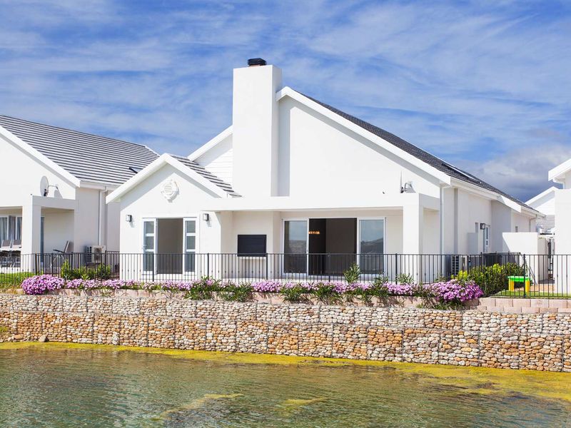 Rear view of a modern white house featuring a prominent chimney, sliding glass doors, and a gabion retaining wall overlooking a water feature.