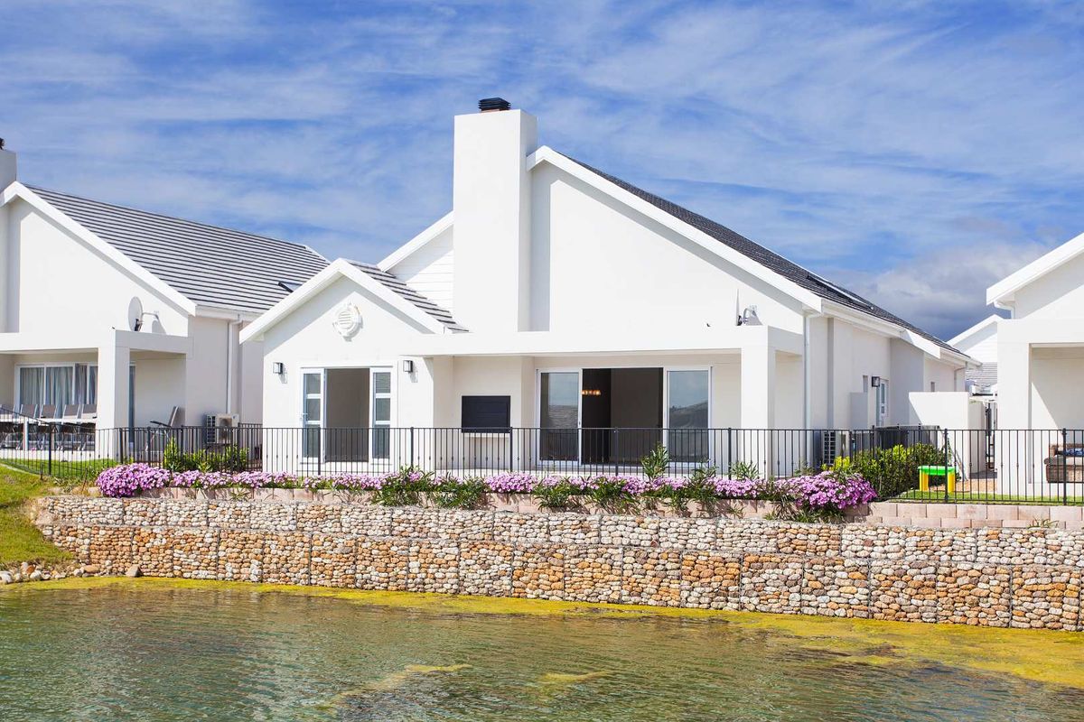 Rear view of a modern white house featuring a prominent chimney, sliding glass doors, and a gabion retaining wall overlooking a water feature.