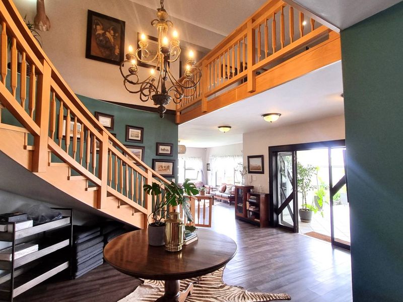 Spacious double-volume entrance hall featuring a wooden staircase, mezzanine landing, elegant chandelier, and direct access to the outdoor patio through sliding glass doors.