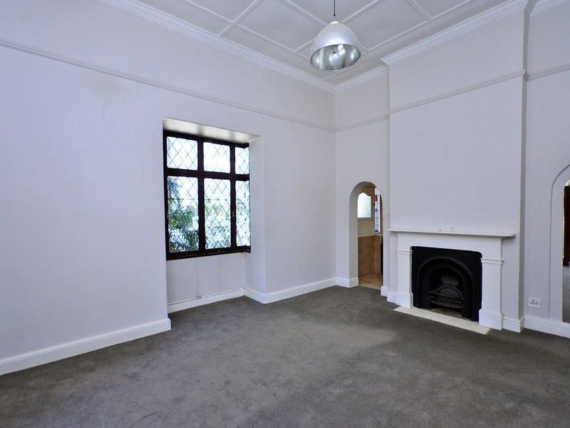 A bright, empty living room featuring a classic fireplace with mantelpiece, leaded glass windows, and elegant coved ceilings.