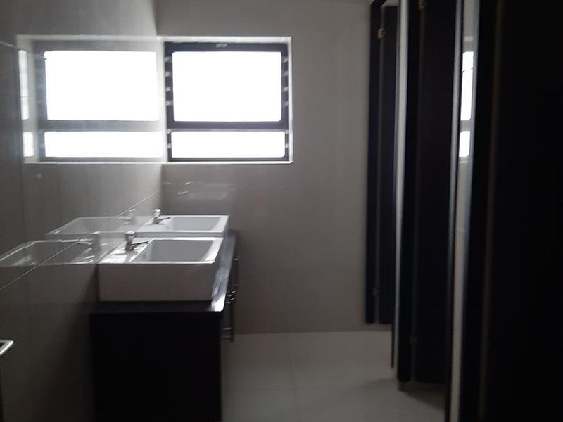 A clean, modern industrial-style communal bathroom featuring a double vanity, tiled finishes, and multiple partitioned toilet cubicles.