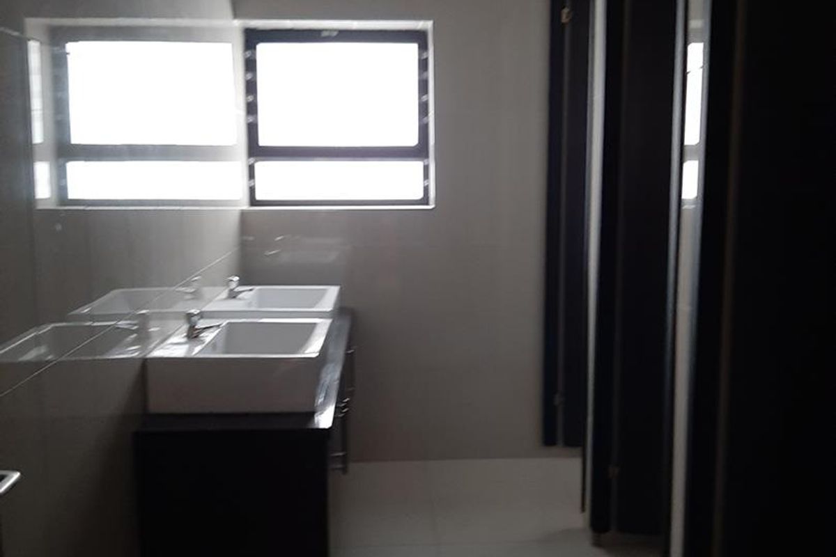 A clean, modern industrial-style communal bathroom featuring a double vanity, tiled finishes, and multiple partitioned toilet cubicles.