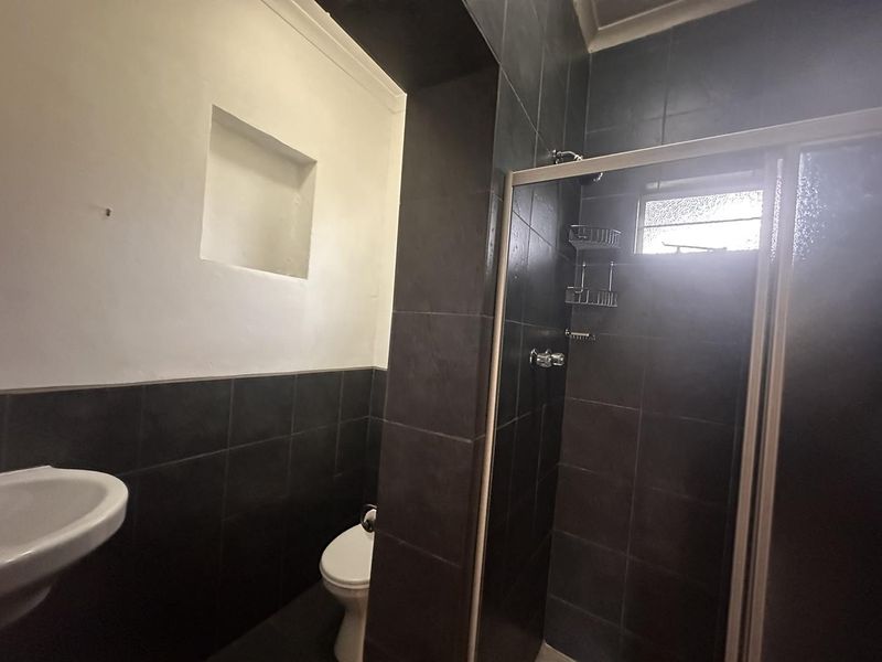 A functional bathroom featuring dark tiled walls, a shower enclosure, a wall-mounted basin, and a toilet with a recessed wall niche.
