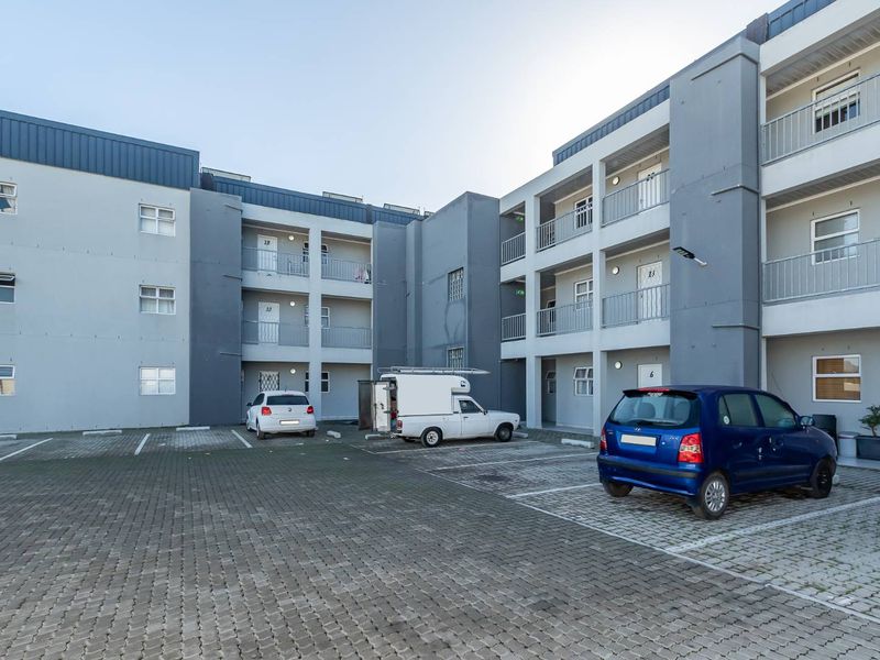 Exterior view of a multi-story apartment complex featuring a paved parking area and building facade with balcony access.