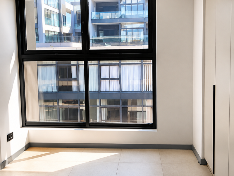 A bright, modern, and empty bedroom featuring large format floor tiles, built-in cupboards, and large black-framed windows with an urban view.
