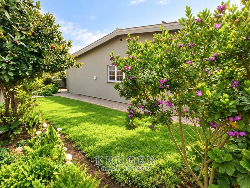 A well-maintained rear garden featuring a lush lawn, established flowering shrubs, and a paved walkway alongside the exterior wall of the house.