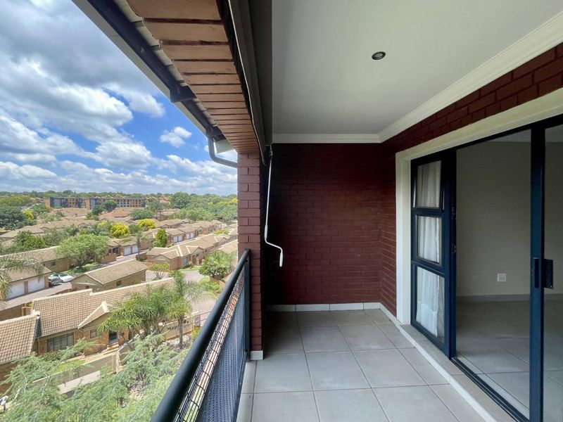 A covered balcony featuring tiled flooring, exposed brick walls, and a metal railing, offering elevated views over the surrounding residential complex.