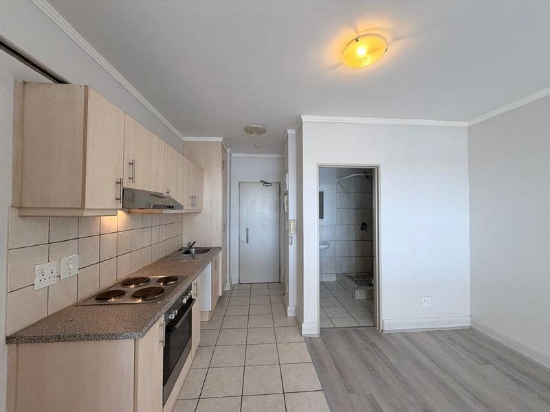 Open-plan kitchen area featuring light wood cabinetry, electric hob, and oven, with tiled and laminate flooring leading to an adjacent bathroom.